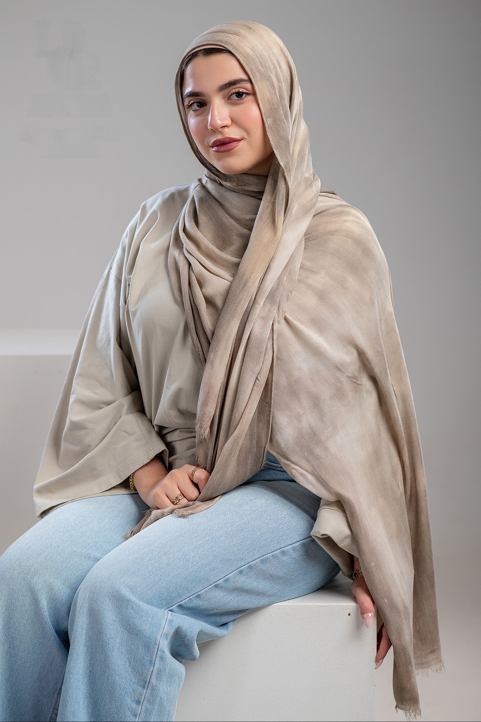 Woman wearing a beige hijab and light blue jeans sitting on a white stool against a gray background